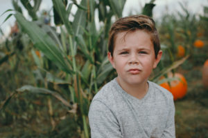 A young boy sits with a very pensive look on his face, among the pumpkins and cornstalks.