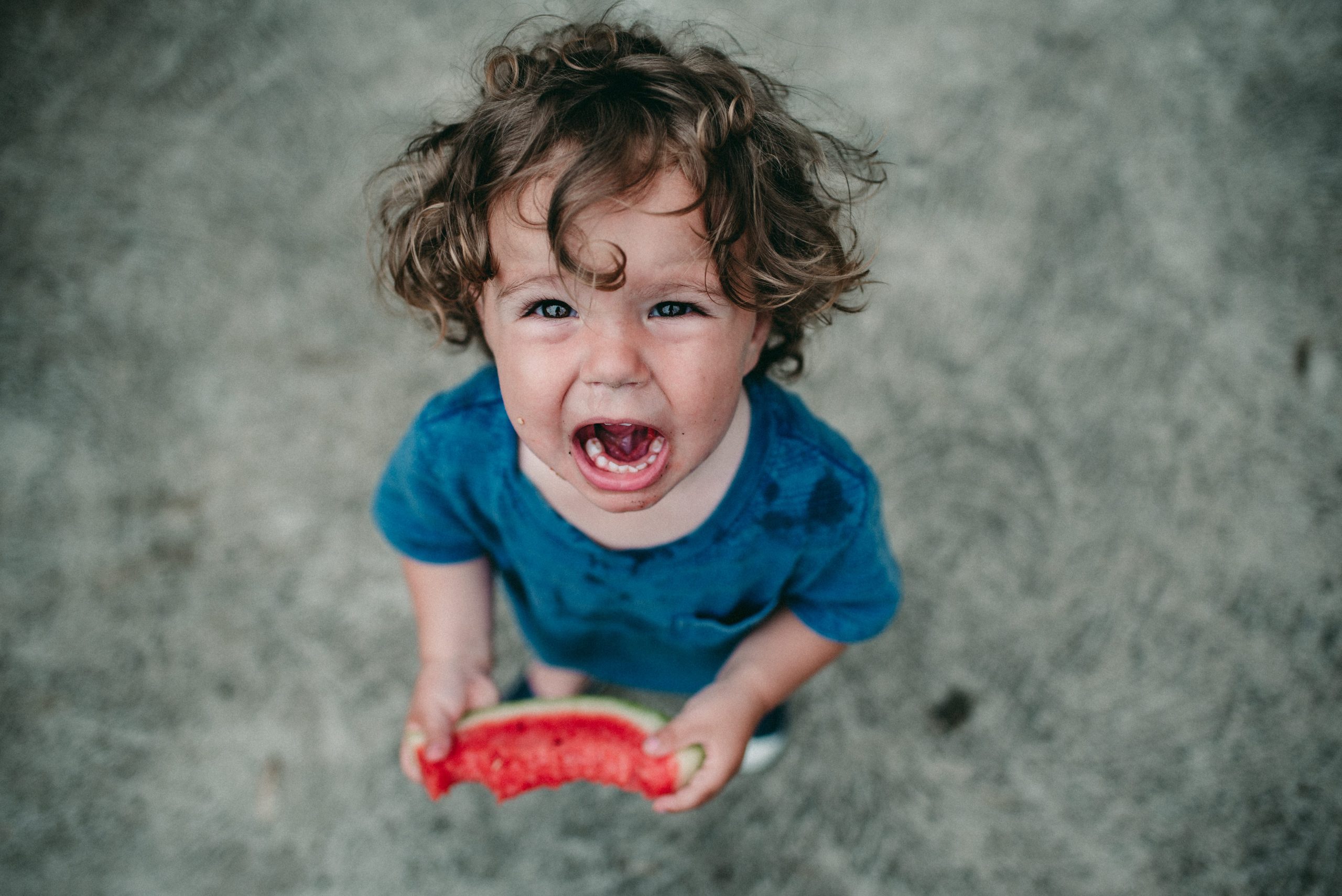 A toddler, shot from above, yells happily with a large piece of watermelon in his hands.