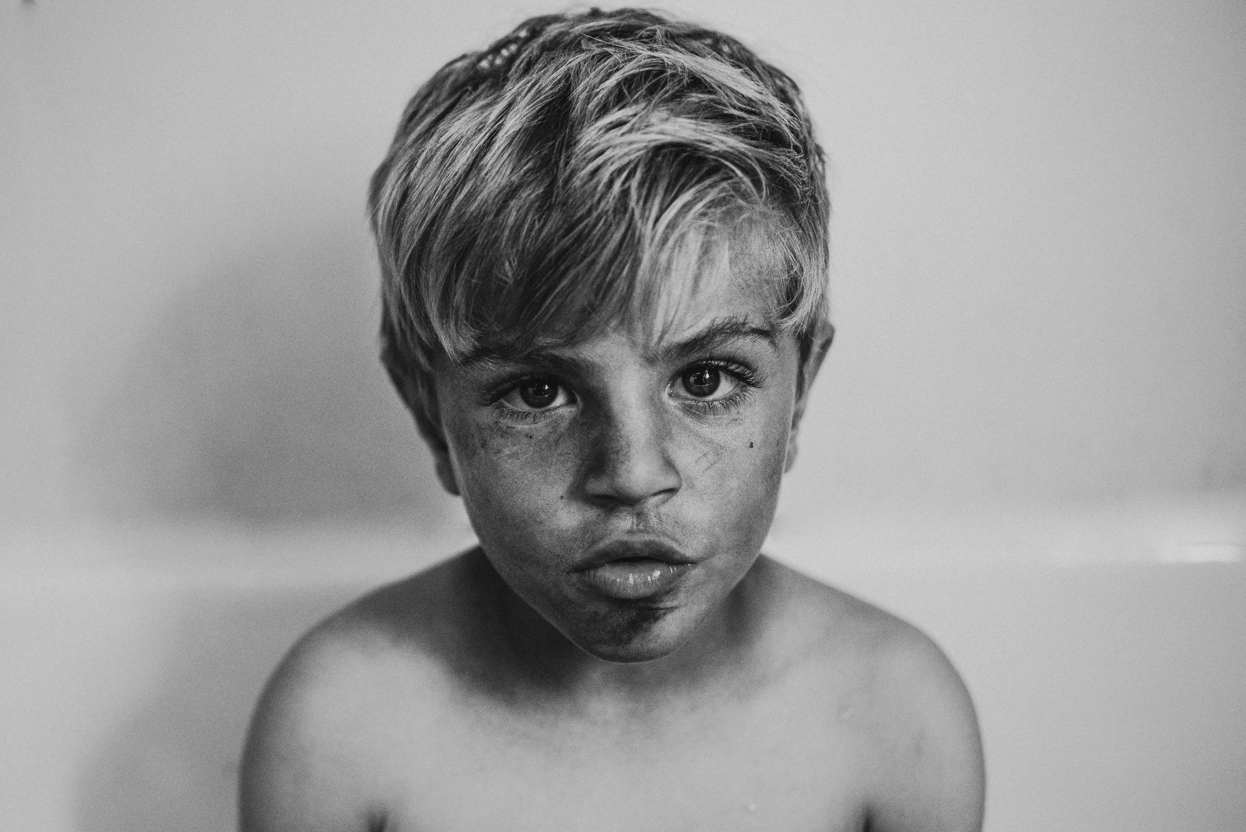 A black and white portrait of a young boy, visible only from the shoulders up, sits in a tub with a very dirty face.