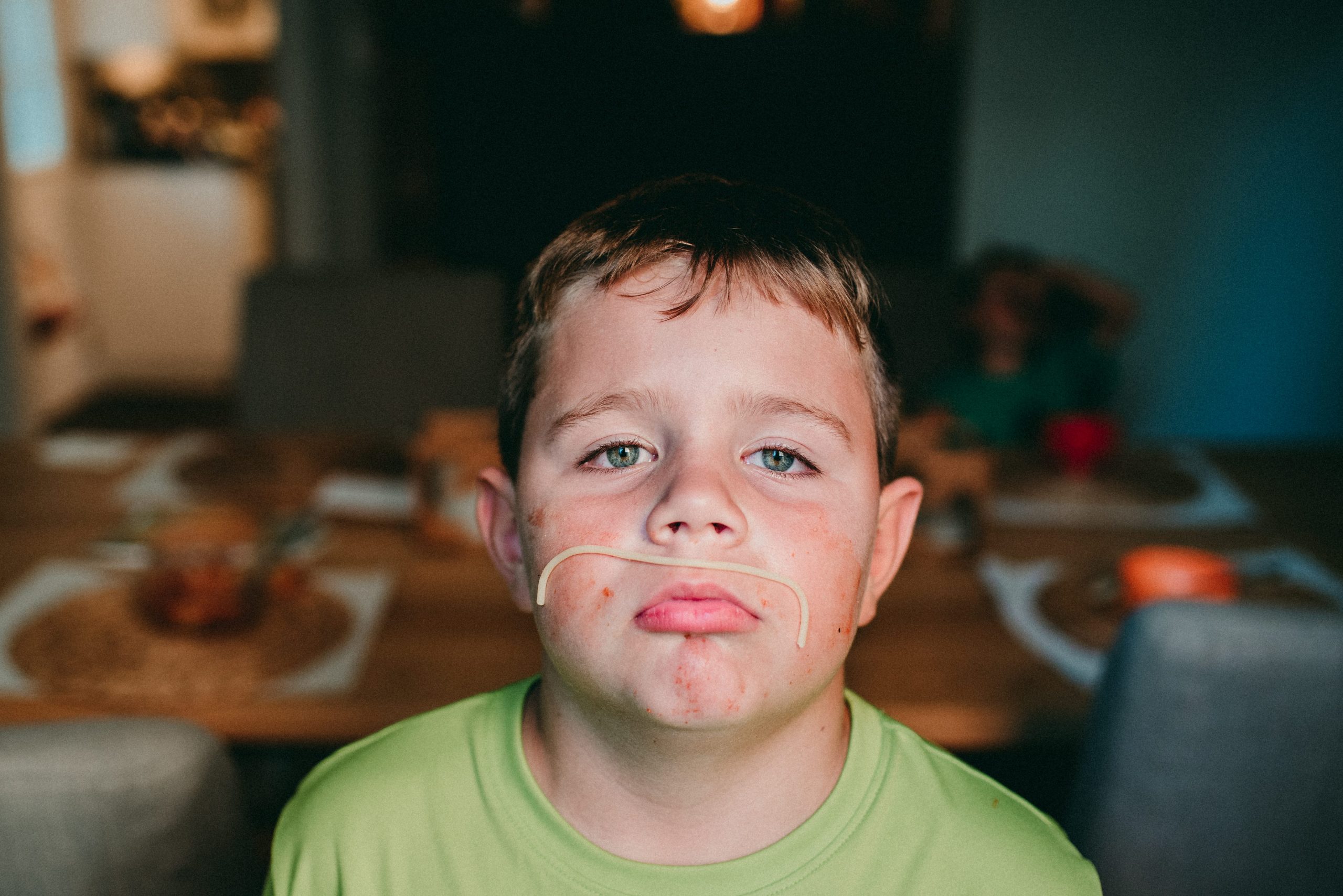 A young boy, lit by window light, poses with a spaghetti noodle mustache.